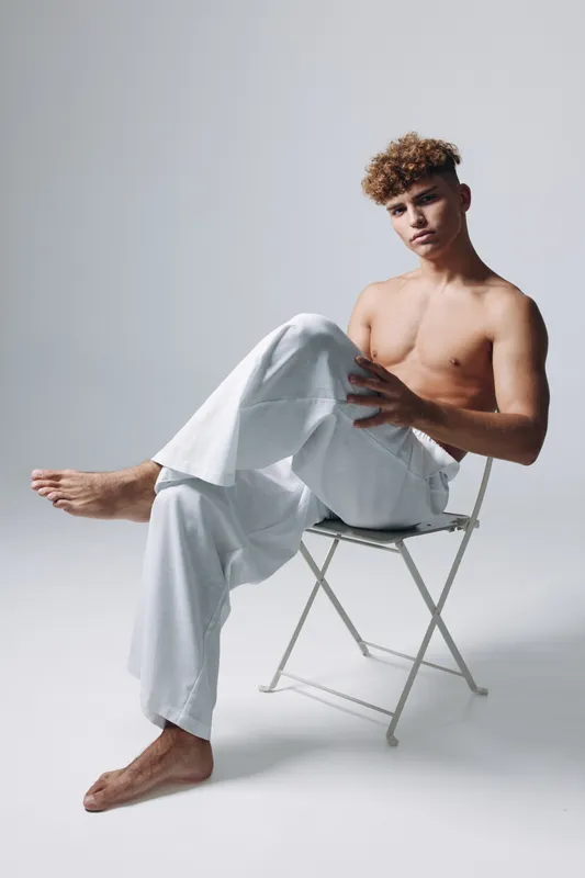 Young man with curly hair sitting shirtless on white chair wearing loose white pants in studio with neutral background. Casual pose and relaxed expression for fashion or lifestyle.