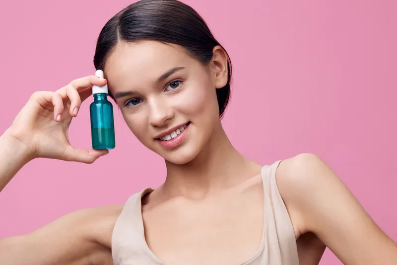 Young woman holding green serum bottle against pink background, promoting skincare and beauty routines for healthy skin and cosmetics