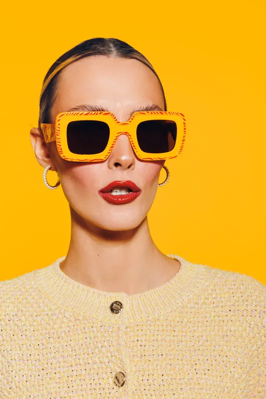 Stylish woman with orange sunglasses and red lipstick poses on bright yellow background. Fashion portrait with trendy accessories and bold makeup. Concept of summer style and confidence.