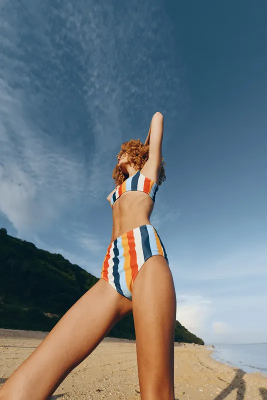 Woman in striped swimsuit stands on a sunny beach, arms raised toward the sky, looking at the ocean. Summer vacation mood with clear blue sky, shoreline, outdoor lifestyle summer feeling.