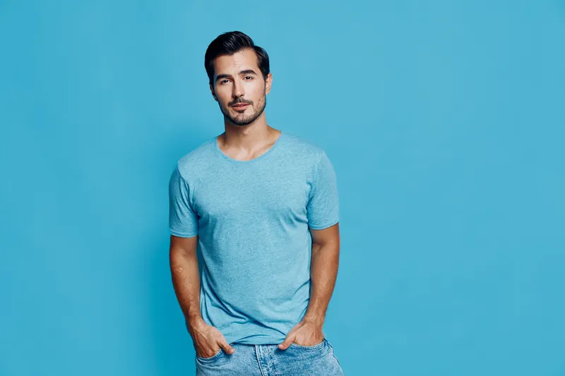 Stylish young smiling man casually dressed in a light blue t-shirt posing confidently against a vibrant blue backdrop, embodying a relaxed lifestyle concept.