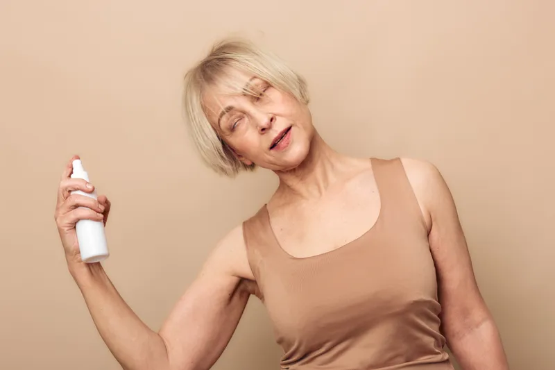 Mature woman with short blonde hair sprays facial mist on her face with closed eyes. Nude background and top wear. Concept for skincare, hydration, and self-care routine for aging skin.
