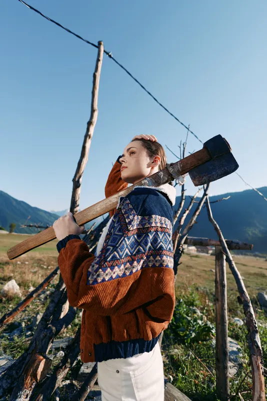 Woman with axe outdoors in mountains wearing patterned sweater in rural setting, portrait by wooden fence and field, standing with thoughtful gaze during sunny autumn day.