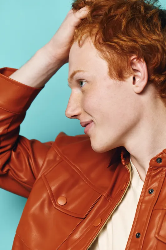 Young man with red hair wearing brown leather jacket posing sideways with hand on head against blue background in studio. Profile portrait of smiling male model.