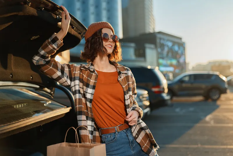 Woman shopping by car trunk in a sunlit parking lot, holding hat and sunglasses, candid lifestyle portrait with golden hour glow, authenticity and mindful living mood.