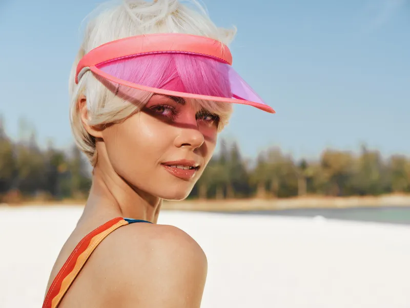 Close-up of a stylish woman at the sunlit beach wearing a pink translucent visor, warm skin glow, and bold makeup, capturing carefree summer mood and playful fashion.