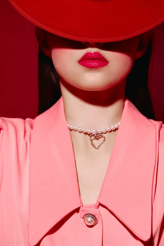 Close-up portrait of a fashionable woman wearing a red hat covering eyes, vibrant pink lips, coral jacket, and delicate pearl necklace with heart pendant. Creative beauty shot.
