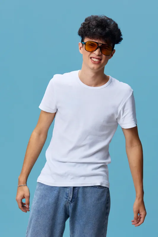 Young man wearing casual white t-shirt and blue jeans smiles confidently against blue background, sporting stylish sunglasses, curly hair, and relaxed pose.