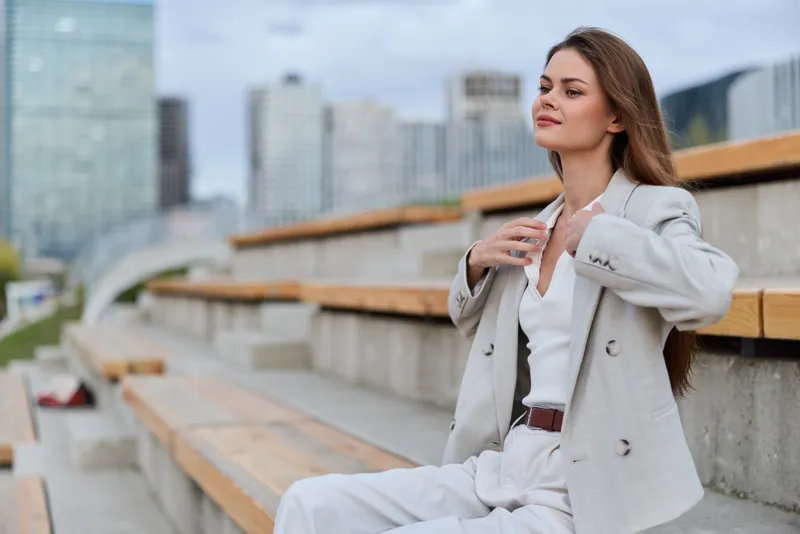 Confident woman in beige business suit sitting outdoors on urban benches with modern city background, calm expression and professional style in spring season