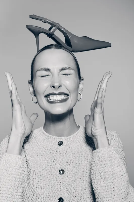 A smiling Caucasian woman with light skin holds black high heel shoe on head, eyes closed and hands raised. Concept of fashion, joy, and playful mood in studio lighting.