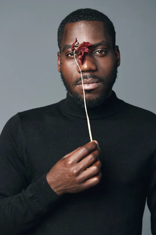 African man with dark skin holding dried flower in front of face in creative conceptual visual style. Portrait with black turtleneck and neutral background for artistic expression and emotion.