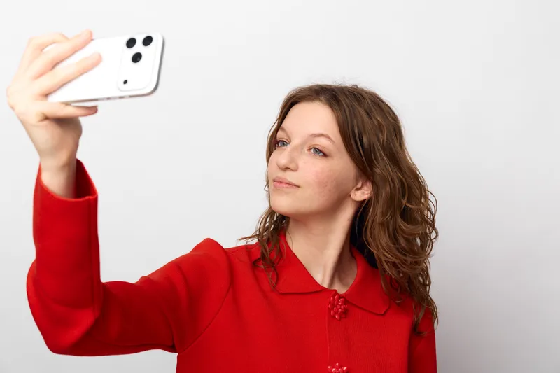 Young woman with curly hair wearing red sweater takes selfie with smartphone on white background. Concept of social media, communication, and modern lifestyle technology.