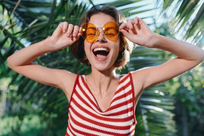 A cheerful young woman wearing a red and white striped top and round orange sunglasses smiles broadly outdoors, enjoying sunny weather in a lush tropical garden with vibrant light.