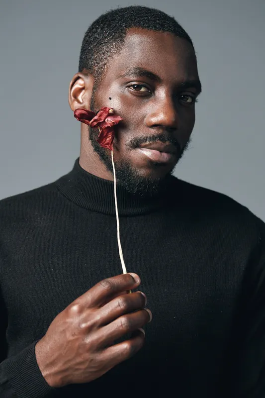 African man with dark skin holding dried red flower near face in creative conceptual visual style wearing black turtleneck sweater on gray background. Portrait with artistic expression and mood.