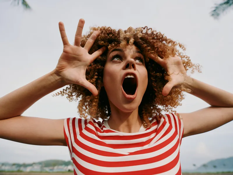 Energetic woman with curly hair wearing a red and white striped shirt shouts outdoors, hands raised near her face, conveying surprise, excitement, and bold movement.