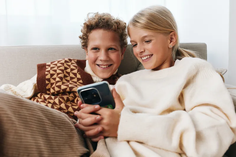 Authentic scene showing children at home, cozy and real, as they share a smile while using a mobile device; play and learning blend in a warm lifestyle moment.
