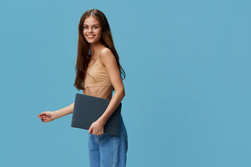 smiling woman holding laptop, wearing casual beige top and blue jeans, standing against solid blue background with long straight hair and cheerful expression