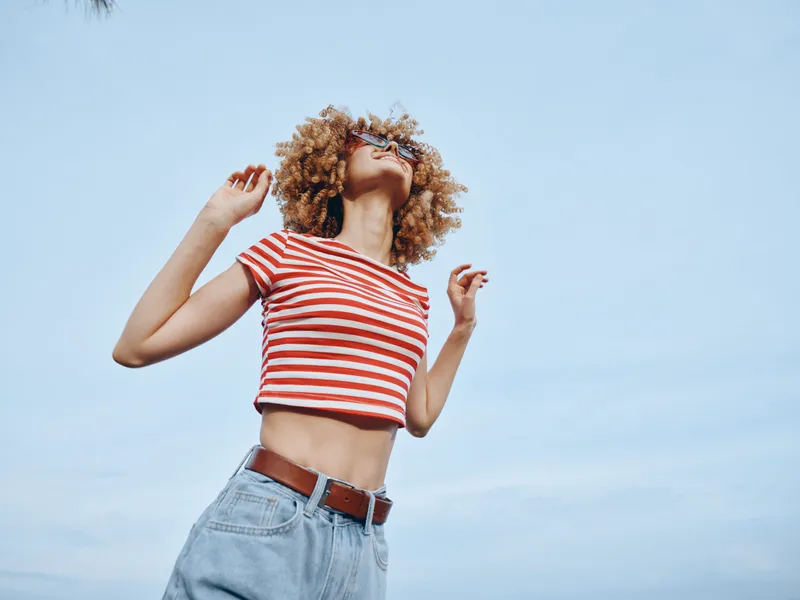 Energetic portrait of a stylish youth outdoors, sunlit sky, striped crop top and denim jeans, carefree pose, bright colors, casual fashion, playful mood, confident stance against clear backdrop.