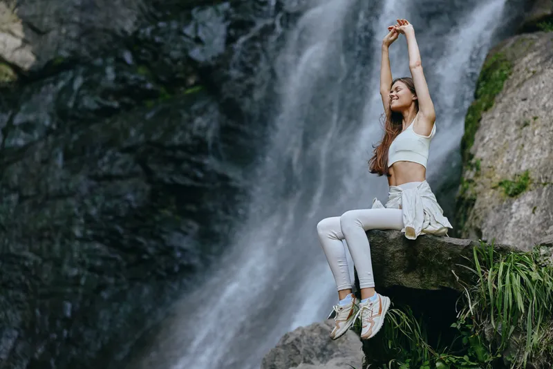 young woman sitting near waterfall, wearing white sportswear, stretching arms, outdoor nature setting, healthy lifestyle and relaxation