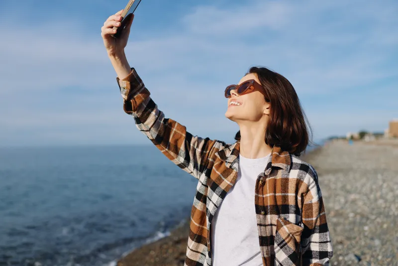 Woman by the ocean taking a selfie with her phone, wearing a plaid shirt and sunglasses, smiling as the sea breeze brushes the beach under a clear blue sky