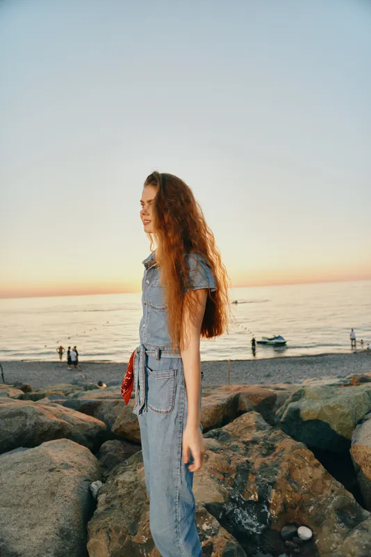 lifestyle woman in modern boho-western denim street style stands on rocky beach at sunset with film color tones and relaxed mood