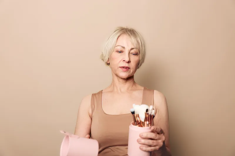 Blonde woman in beige top holds pink container with makeup brushes, looking down with calm expression. Neutral background creates soft tone for beauty, cosmetics, and skincare concept.