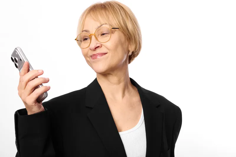 Smiling older woman in glasses holding a smartphone, portrait on white background, black blazer, business communication, technology and connection, calm confidence