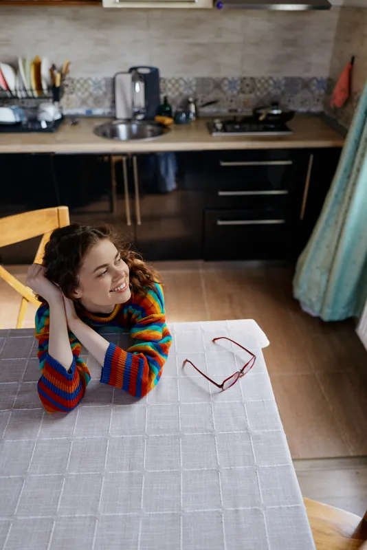 A relaxed woman sits at a kitchen table with her head in her hands, smiling softly. A bright home scene, colorful striped sweater, and glasses on the table create a cozy lifestyle moment.