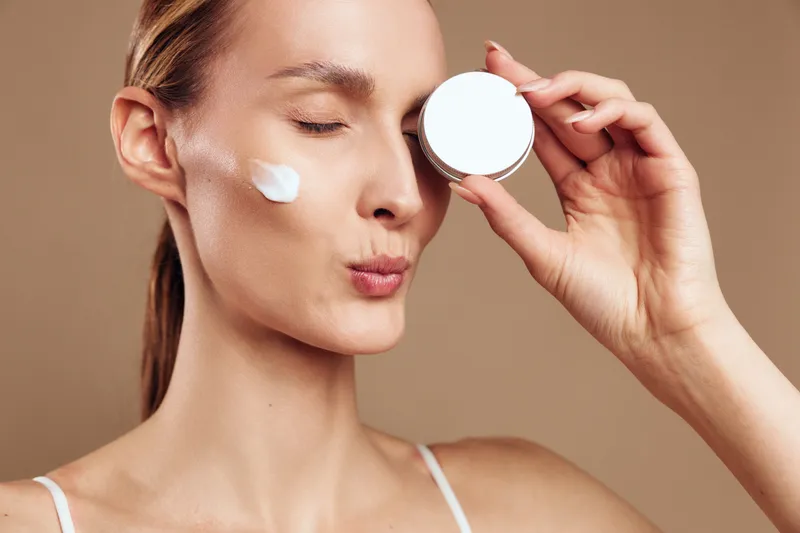 Young woman with smooth skin applying white face cream on cheek while holding cosmetic jar close to face on beige background. Concept of skincare, beauty, self-care, and wellness.