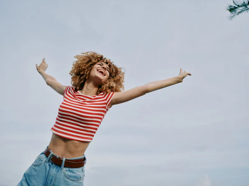 Joyful young woman outdoors with arms outstretched, wearing a red and white striped crop top and blue jeans, capturing carefree happiness against a bright sky.