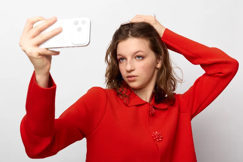 Young woman in red sweater takes selfie with smartphone, posing with hand in hair against white background. Concept of social media, self-portrait, and casual lifestyle.