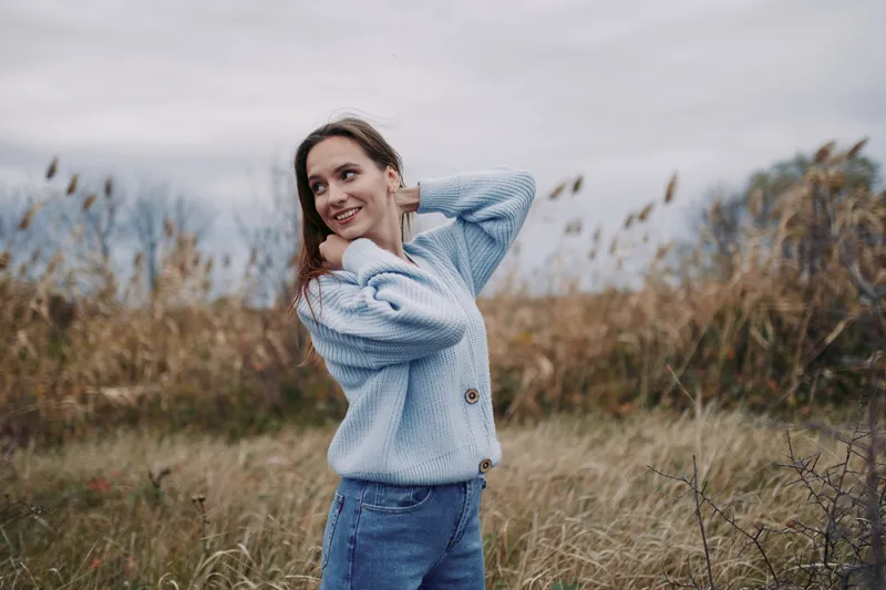 Portrait of a young woman in a cozy blue knit sweater standing in a windswept meadow, candid smile and relaxed posture conveying authentic emotion and believable everyday charm.