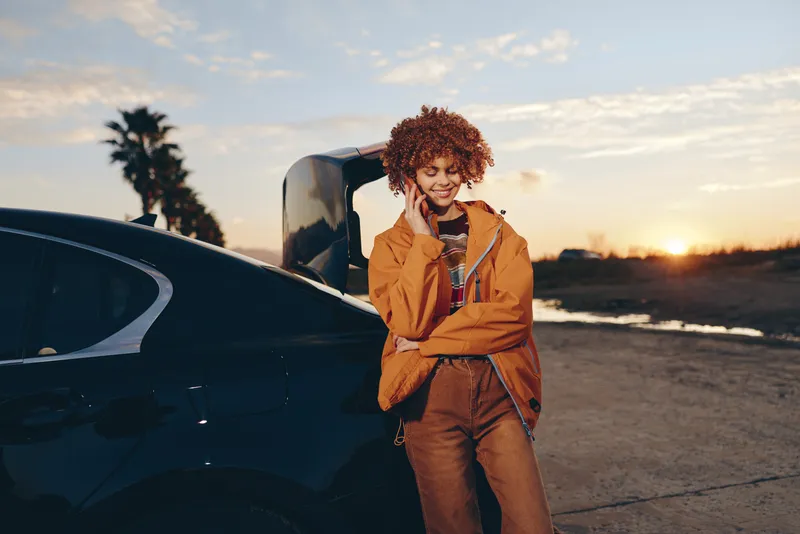 Woman lifestyle outdoors near a car at sunset, curly hair woman in orange jacket smiling and laughing, relaxed pose by open door, warm light, coastal road background