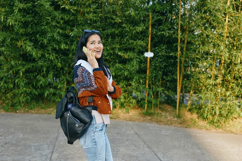 Smiling woman stands outdoors talking on a cell phone, wearing a brown jacket, cozy sweater, and light jeans with a backpack beside a green hedge on a bright day