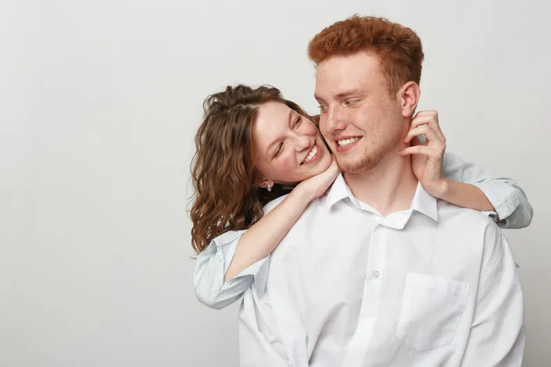 Happy young couple in casual white shirts embraces and smiles, showing affection and joy. Studio portrait on light background captures love, friendship, and positive emotions between two people.