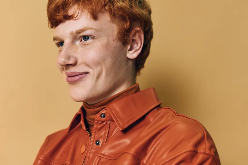 Young man with red hair and freckles wearing orange leather jacket and turtleneck smiling with confidence against beige studio background. Portrait of casual style.