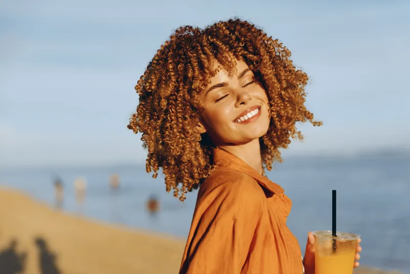 Smiling young woman with curly hair portrait in brown jacket enjoys sunny beach by the ocean, holding a fresh drink cup, carefree summer travel mood with soft coastal background vacation