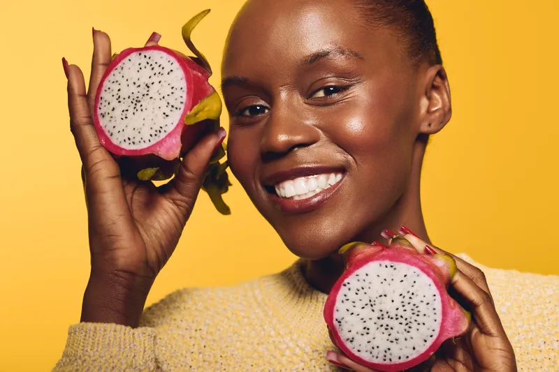 Smiling dark skinned African woman holds two halves of dragon fruit near her face. Bright yellow background and warm lighting highlight healthy skin and fresh tropical fruit concept.