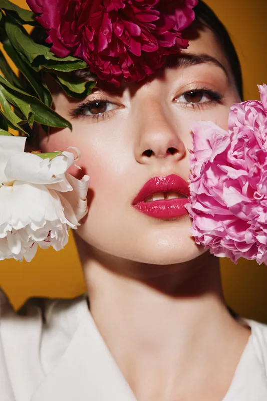 Close-up portrait of woman with vibrant floral makeup look holding colorful flowers near face. Creative beauty shot on orange background. Fashion and cosmetic concept.