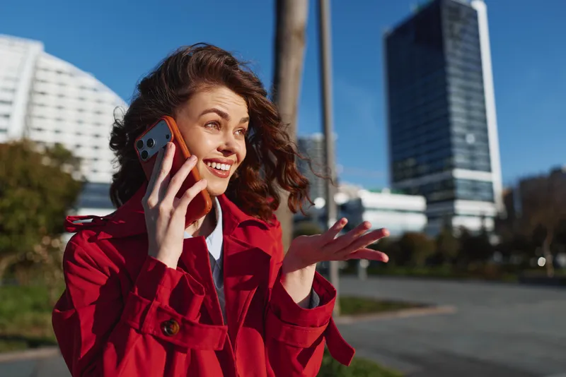 Young woman in red jacket talks on smartphone outdoors in an urban environment with modern buildings under clear blue sky.