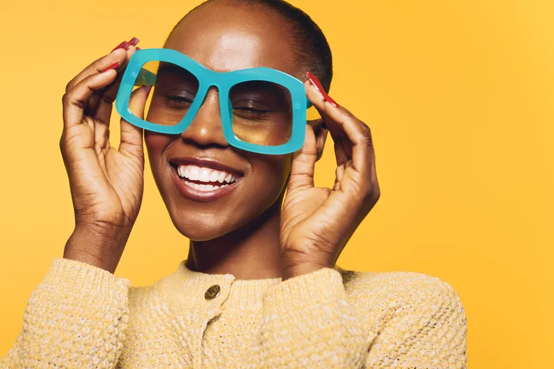 Happy African woman with dark skin wearing oversized blue glasses and yellow sweater smiles with closed eyes on yellow background. Concept of joy, fashion, and positive mood.