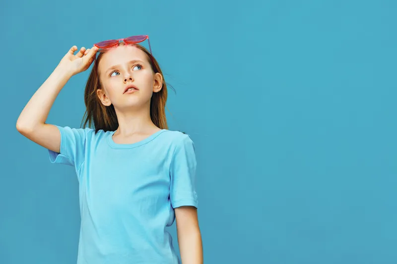 Cute girl in a blue t shirt and pink sunglasses looking up against a blue background, portraying curiosity and playfulness during summer