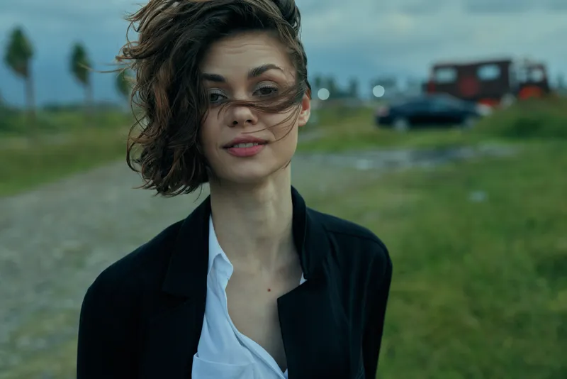 Windy outdoor shot of a woman in a black jacket and white shirt, windblown hair, moody weather, grassy field with distant car in the background