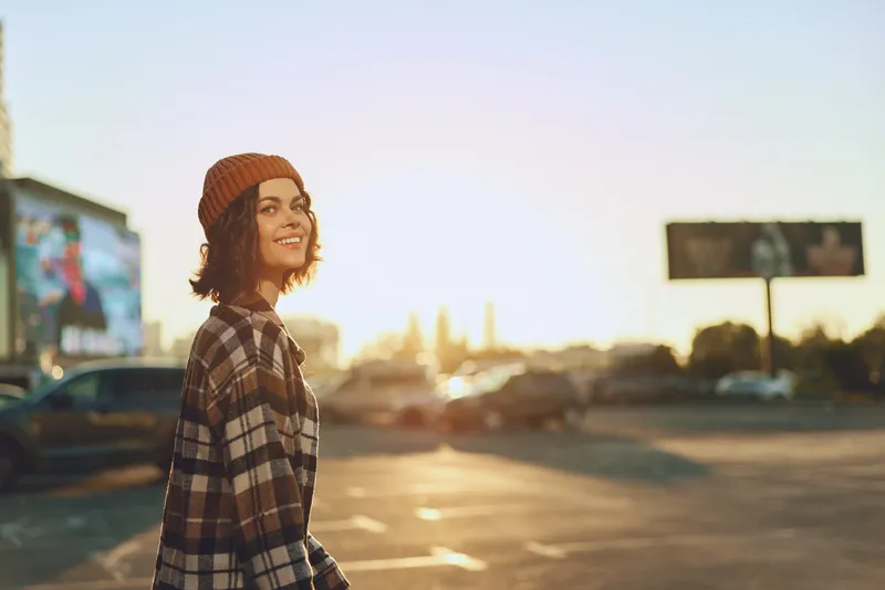 Woman with beanie and plaid shirt smiling in urban parking at golden sunset, candid lifestyle portrait conveying authenticity, mindful living and emotional storytelling with warm golden hour glow.