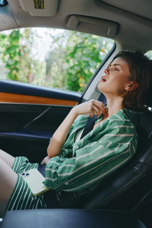 A young person sings with mouth open in the car, wearing a green striped shirt, seatbelt fastened, sunlight streaming through the window.