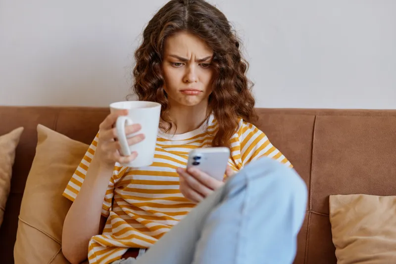 Woman lifestyle at home sits on couch with striped top holding a mug and looking at smartphone, casual cozy living room, thoughtful moment, everyday solitude