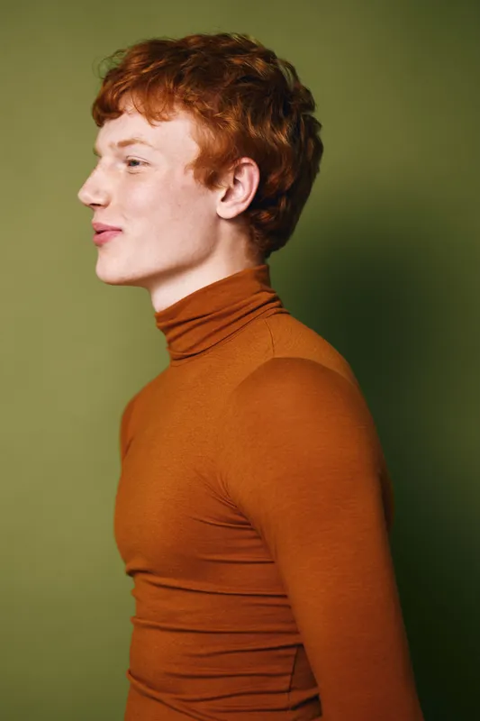 Young man with red hair and pale skin wearing orange turtleneck sweater posing in profile against green background. Stylish casual portrait in studio with neutral expression and soft lighting.