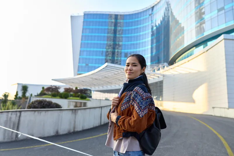 A woman with backpack stands beside a curved glass building in an urban setting, showcasing modern fashion, daylight mood and travel vibe, casual outerwear, confident pose, street style, contemporary