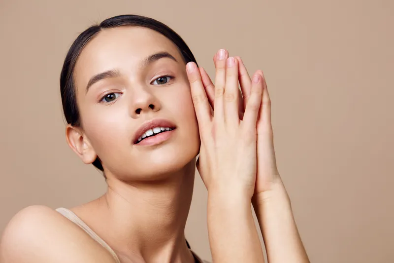 Natural beauty portrait of a young woman with clear skin, soft smile, and long hair against a neutral background, emphasizing self care and confidence
