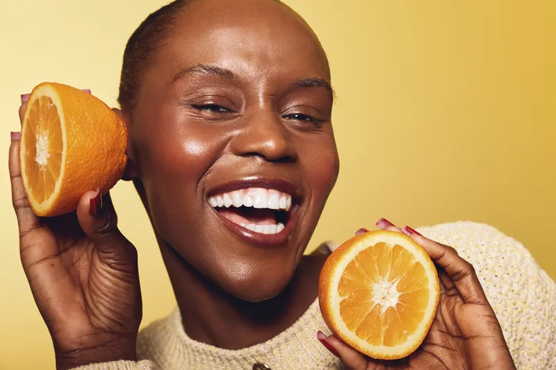 Happy African woman with dark skin holds two halves of fresh orange, smiling with teeth on yellow background. Concept of healthy lifestyle, vitamin C, wellness, and natural beauty.